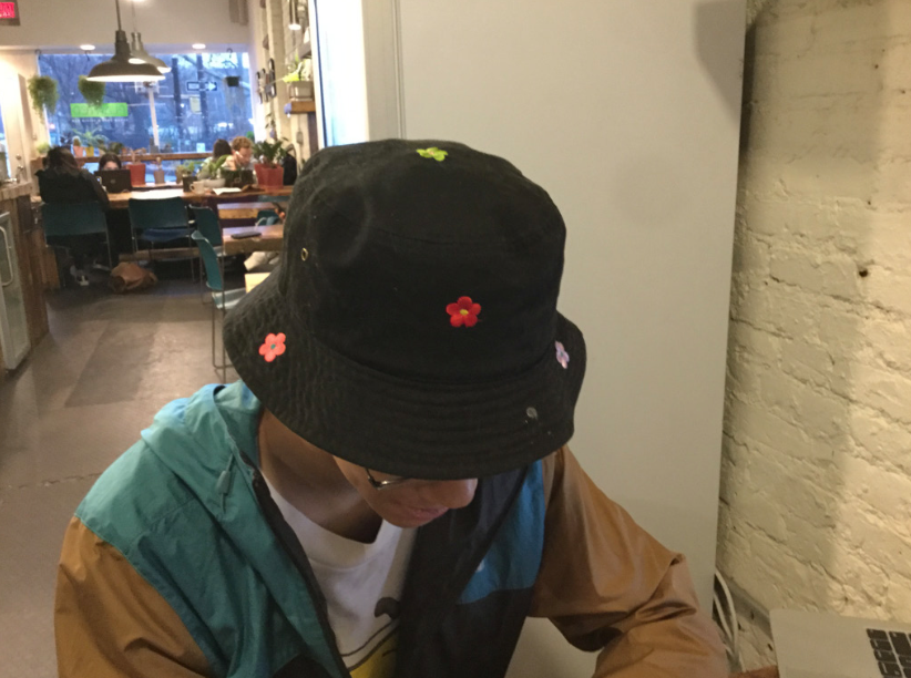 Taichi is sitting in a cafe, looking downward at his phone. He wears a black bucket hat sprinkled with embroidered flowers of various bright colors.
